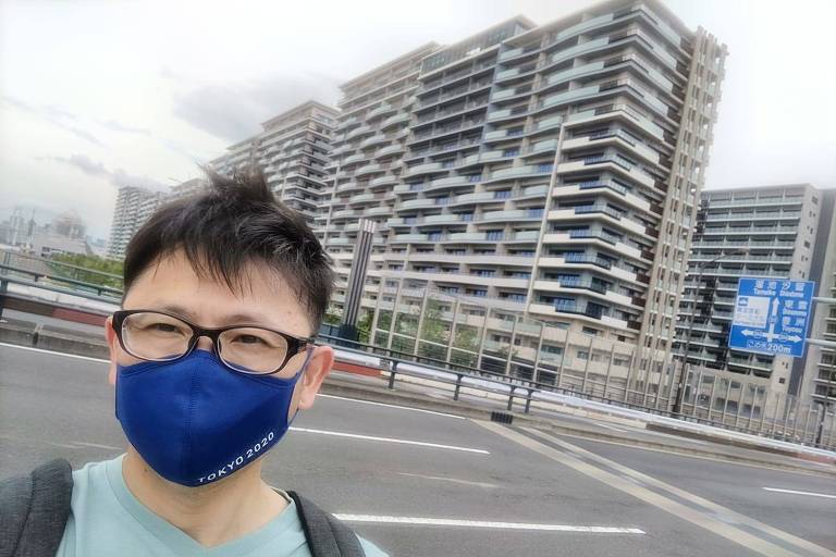 Homem de máscara tira selfie em frente a prédios da Vila Olímpica