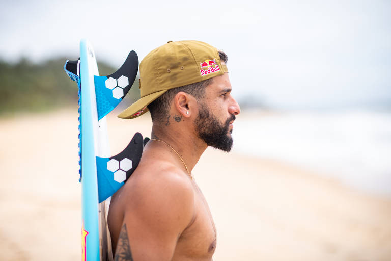 Retrato de Italo Ferreira, de lado, com a prancha nas costas, na praia de Baia Formosa, no Rio Grande do Norte