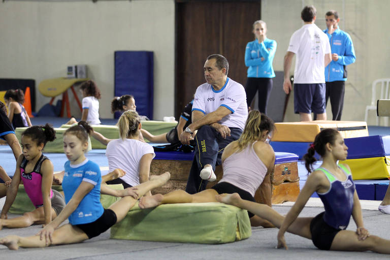 O técnico Oleg Ostapenko, ao centro da foto, comanda treino da seleção brasileira de ginástica artística. Ao redor do treinador estão várias ginastas alongando as pernas
