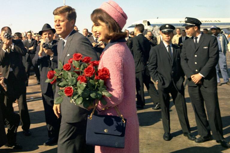 A imagem mostra John F. Kennedy e Jacqueline Kennedy sendo recebidos em um aeroporto. Jacqueline está vestida com um vestido rosa e um chapéu combinando， segurando um buquê de rosas vermelhas. JFK está ao seu lado， usando um terno escuro. Eles estão cercados por uma multidão de pessoas， incluindo fotógrafos e membros da equipe de segurança. Ao fundo， é possível ver a asa de um avião e outros passageiros.