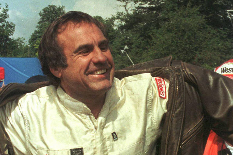 Carlos Reutemann, ex-piloto de Fórmula 1, sorri. Ele está vestido com roupa de piloto e coloca uma jaqueta