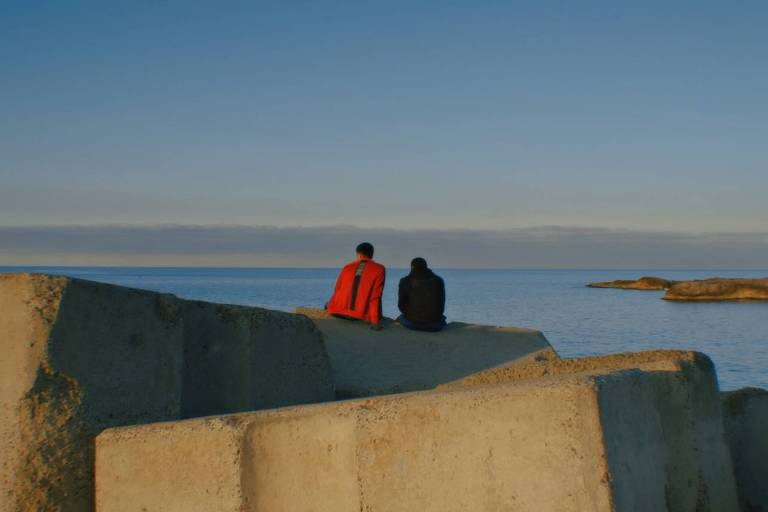 Duas pessoas aparecem sentadas em pedras e, ao fundo, vê-se o mar