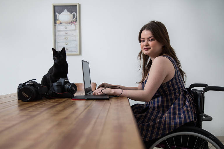Mulher branca, de cabelos longos escuros e vestido xadrez, em cadeira de rodas, à mesa onde estão um computador, equipamentos fotográficos e um gato preto