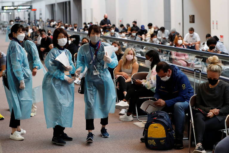 Pessoas sentadas em fila e funcionários vestidos com roupas de proteção em saguão do aeroporto
