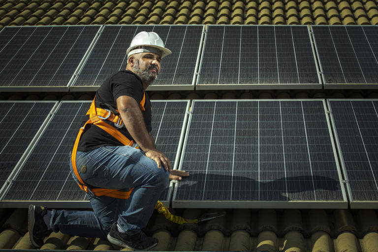 Homem de capacete e equipamento de segurança instalando painéis de energia solar, que são quadrados escuros, em um telhado