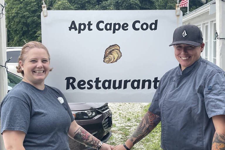 Brandi Felt Castellano (esquerda) e Regina Felt Castellano (direita), posam para foto em frente à placa de seu restaurante, que é branca com letras pretas. Elas vestem camisetas cinza. Regina usa um boné. 
