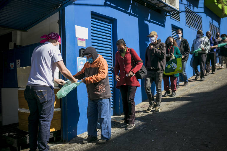 A imagem mostra uma fila de pessoas aguardando para receber alimentos em um local com paredes azuis. Um homem, usando uma camiseta branca e um lenço rosa na cabeça, está entregando um pacote verde a um homem idoso, que usa um casaco laranja e um chapéu. Ao fundo, outras pessoas estão na fila, algumas usando máscaras. O ambiente parece ser um espaço comunitário ou de assistência social.