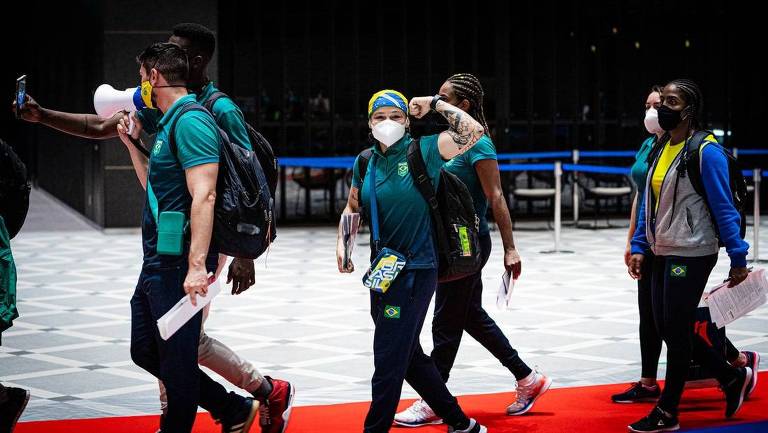 Atletas caminham em saguão do aeroporto; ao centro, Beatriz Ferreira mostra o muque