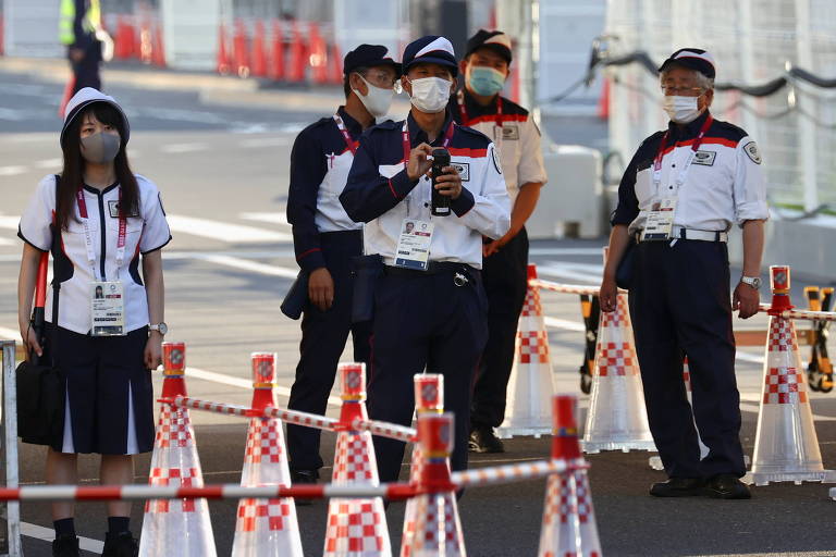 Seguranças e membros do staff trabalham na entrada da Vila Olímpica. Todos estão uniformizados e com máscaras de proteção