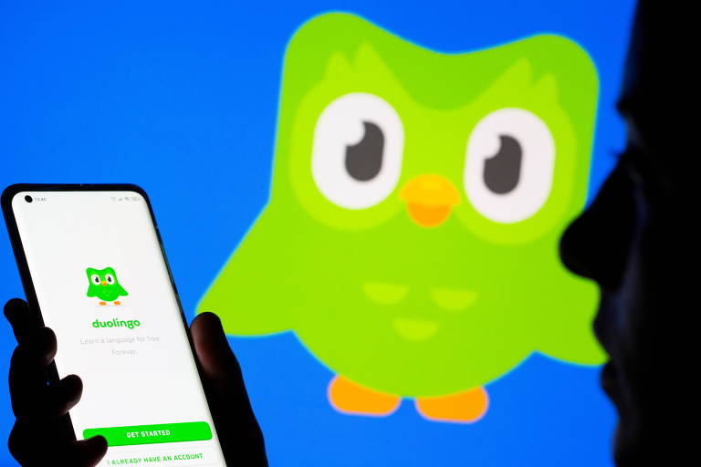 A imagem mostra uma pessoa segurando um smartphone com a tela voltada para a câmera, exibindo o aplicativo Duolingo, caracterizado pelo ícone de uma coruja verde. O fundo azul e o perfil silhueteado da pessoa criam um contraste que destaca o telefone e o aplicativo.
