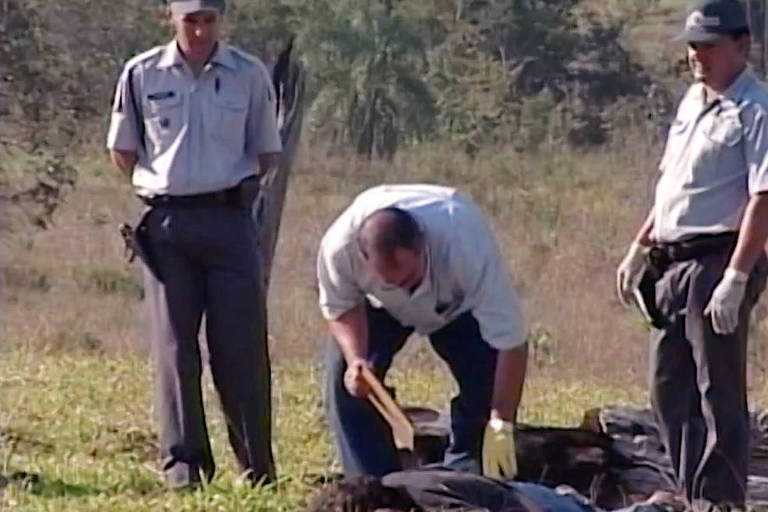 Três policiais perto de corpos em uma área rural 