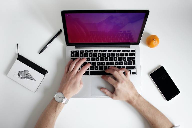 mãos digitam em computador sobre mesa branca. Há ao lado dele um celular, uma fruta, uma caneta e mais um aparelho tecnológico