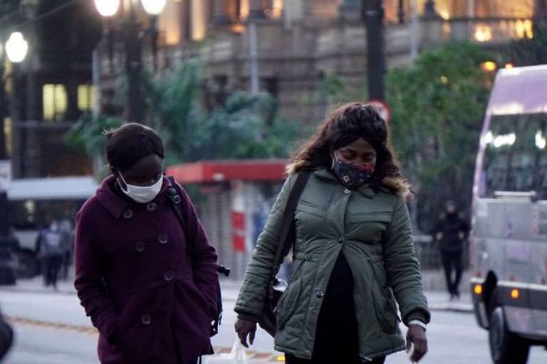 Duas mulheres negras de casaco e máscara caminham por uma rua