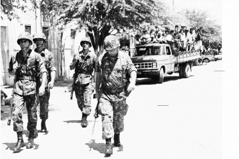 Imagem em preto e branco mostra militares caminhando a frente de um caminhão, de onde descem vários militares
