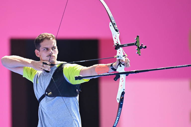 Um atleta está posicionado em uma postura de tiro com arco, segurando um arco com uma flecha pronta para ser disparada. Ele usa uma camiseta cinza e um colete de proteção preto. O fundo é de cor rosa, destacando a ação do atleta.