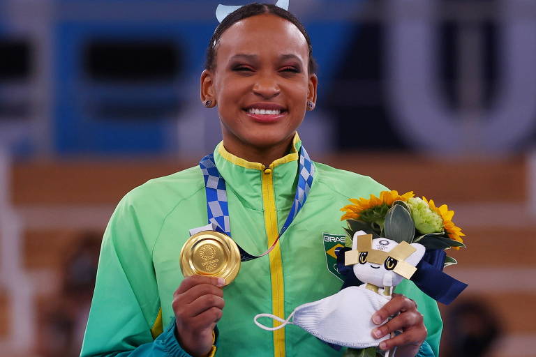 Uma mulher negra com uniforme da seleção brasileira sorri e segura sua medalha