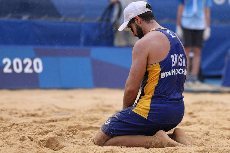 Aloelhado na areia da quadra de vôlei de praia no parque Shiozake, Bruno Schmidt, que fez parceria com Evandro, lamenta a eliminação nas Olimpíadas de Tóquio, nas oitavas de final; ele usa boné branco e uniforme azul e amarelo