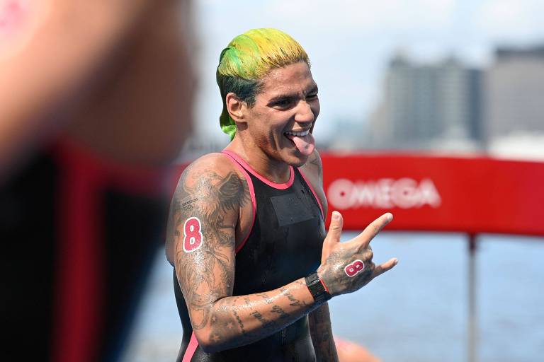 Ana Marcela Cunha, com cabelo pintado de verde e amarelo, mostra a língua e faz um sinal com a mão após sair do mar na prova olímpica
