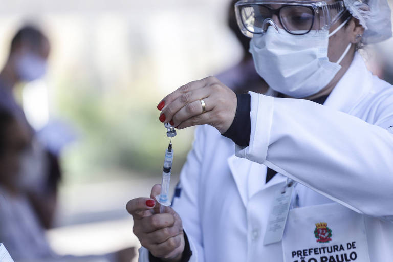 mulher, de jaleco, máscara e touca cirúrgica, prepara vacina