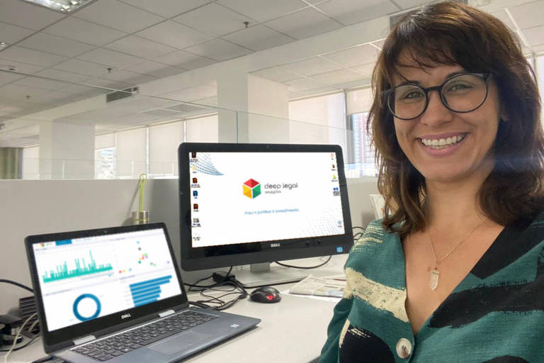 Uma mulher branca e de óculos ao lado de telas de computador