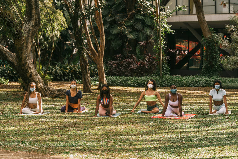 Seis mulheres negras estão sentadas em um tapete de ioga em um gramada. Ao redor, há vegetação.