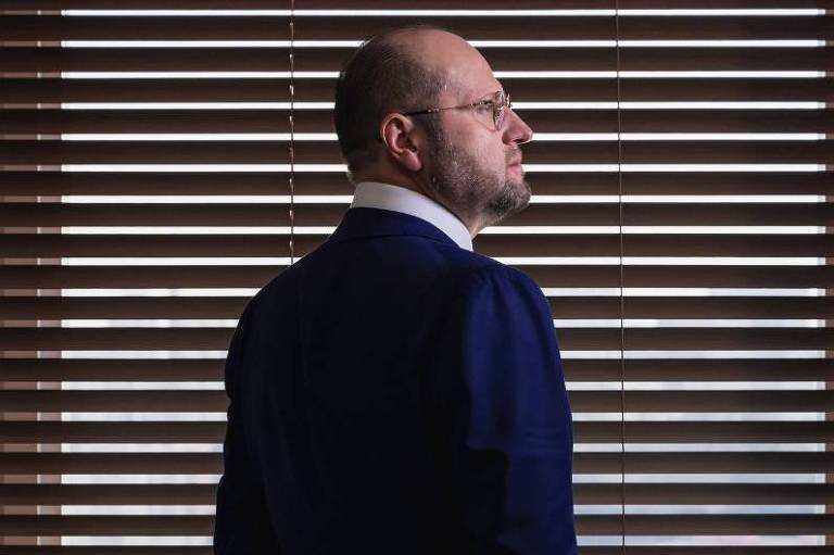 Advogado Nelson Wilians de perfil em frente a uma janela, com terno azul