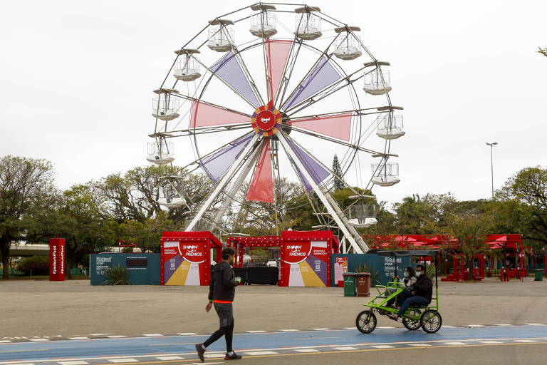 Ao fundo da foto, uma roda gigante de 20 metros de altura com o logo do "iFood" no centro. Em primeiro plano, um casal em um triciclo para a família e uma pessoa andando passam pela ciclovia do parque Ibirapuera.