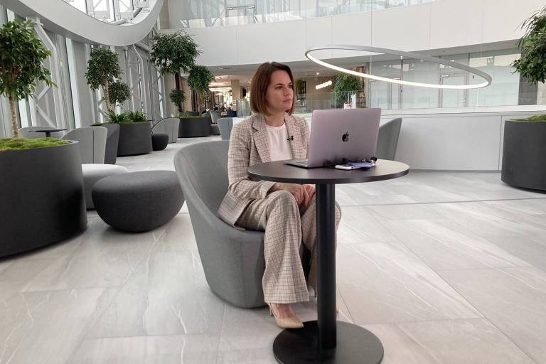 mulher de cabelos castanhos escuros cultos e terno calro, em saguão amplo com grandes janelas, posa em frente a um laptop aberto sobre uma mesa