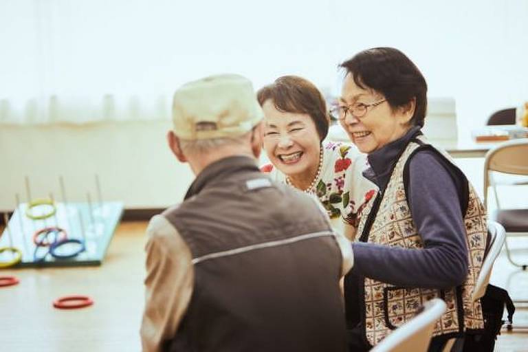 Imagem em primeiro plano mostra três idosos, uma mulher e um homem. Eles estão sentados de frente para uma mesa. As duas mulheres sorriem e o homem está de costas