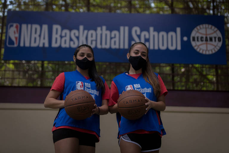 Adolescentes usando máscara de proteção contra a Covid-19 seguram bolas de basquete, em frente ao letreiro do NBA Basketball School