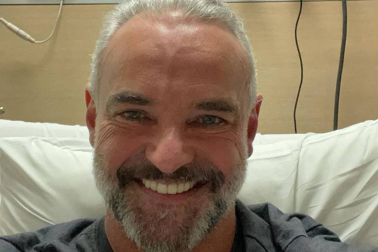 Homem branco com cabelo e barba grisalhos sentado em cama de hospital