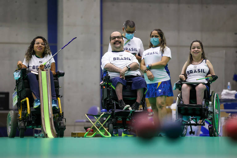 Três atletas em cadeiras de rodas sorriem atrás da pista de bocha