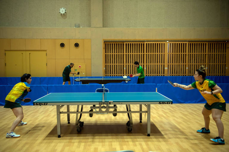 Atletas, cada uma de um lado da mesa, trocam bolas durante o treino