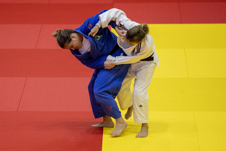 Judocas, uma de azul e outra de branco, seguram os quimonos uma da outra e se movimentam sobre o tatame vermelho e amarelo