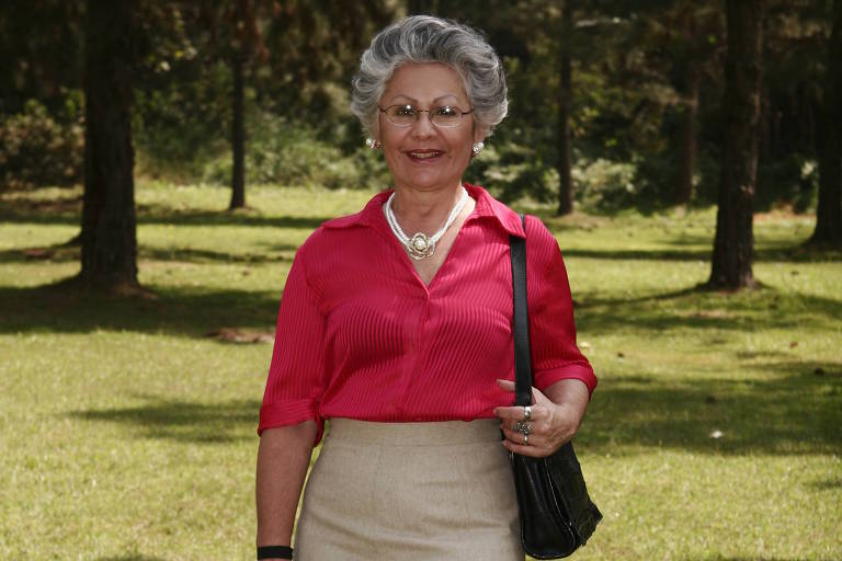 Senhora branca de cabelos grisalhos em um parque vestindo óculos, saia lápis bege, suéter rosa, bolsa preta e sandália de salto preto