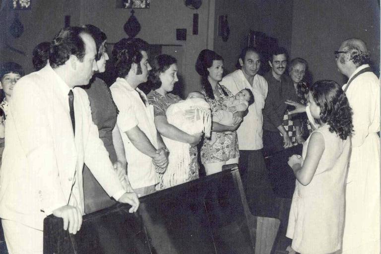 Imagem antiga em preto e branco. Em uma igreja, várias pessoas estão em pé. Ao centro, duas mulheres seguram seus filhos, enquanto um padre os batiza