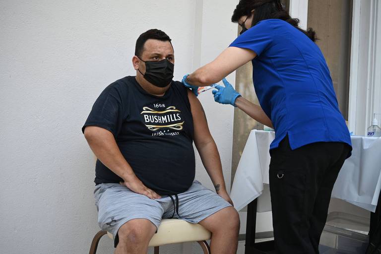 Homem de máscara e camiseta pretas, sentado em cadeira, recebe vacina de profissional de saúde nos Estados Unidos