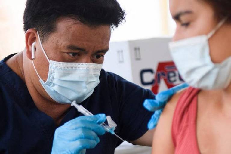 Um profissional da saúde com uma seringa na mão se prepara para aplicar a vacina no braço de uma mulher.​