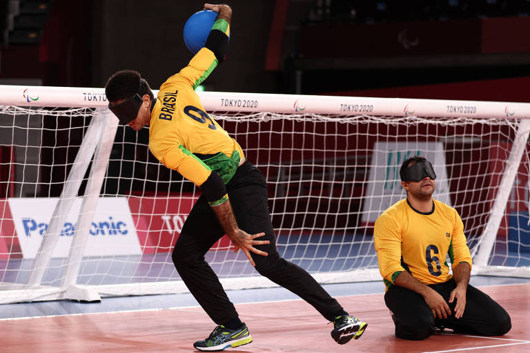 Jogadores com camisetas amarelas e calças escuras; um deles está de joelhos no chão em frente ao gol, e o outro está de pé preparando o lançamento da bola