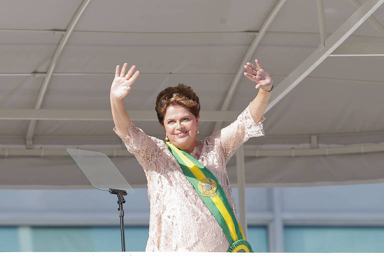 Dilma aparece sorrindo e acenando com suas duas mãos para o alto, acima de sua cabeça. Ela foi fotografada da cintura para cima. Usa um vestido branco e a faixa presidencial em verde e amarelo. 