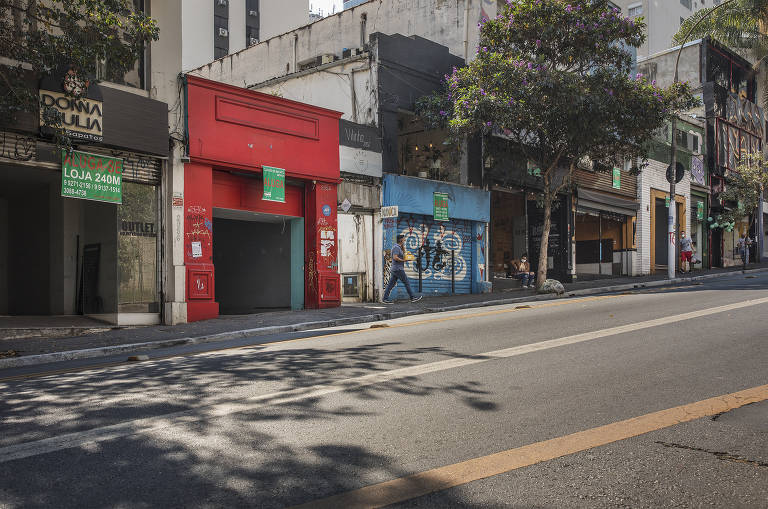 A imagem mostra uma rua com várias fachadas de lojas. À esquerda， há uma loja com uma entrada vermelha e uma placa verde. Ao lado， uma parede azul com grafite. As lojas estão fechadas e a rua está vazia