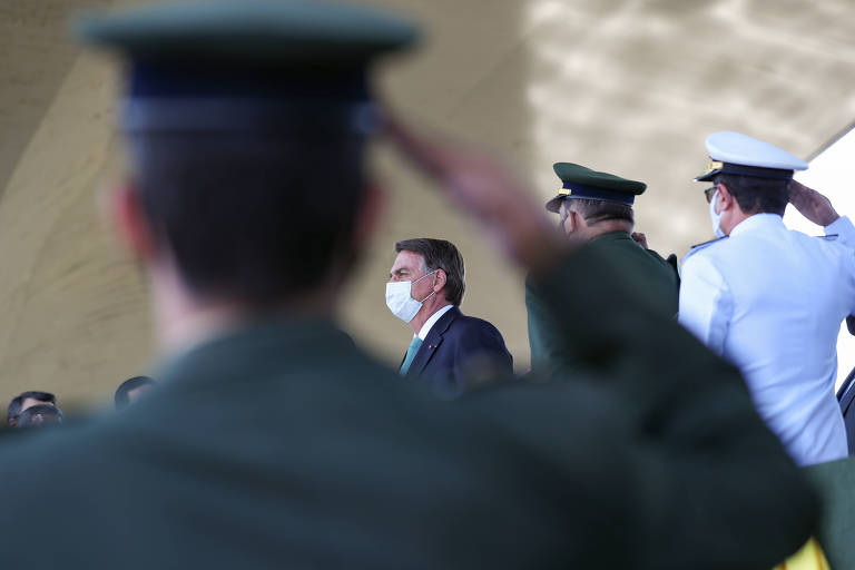 Em primeiro plano, de costas, um militar fardado fazendo continência a Bolsonaro, que aparece no espaço estre o braço e a cabeça do militar. Ao lado dele, outros dois militares fardados