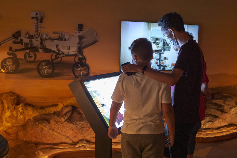 Duas pessoas, um adulto e uma criança, observam a um totem digital da exposição 'Fututo Espacial'. Ao fundo está um cenário alaranjado com rochas no chão e uma réplica de robô espacial