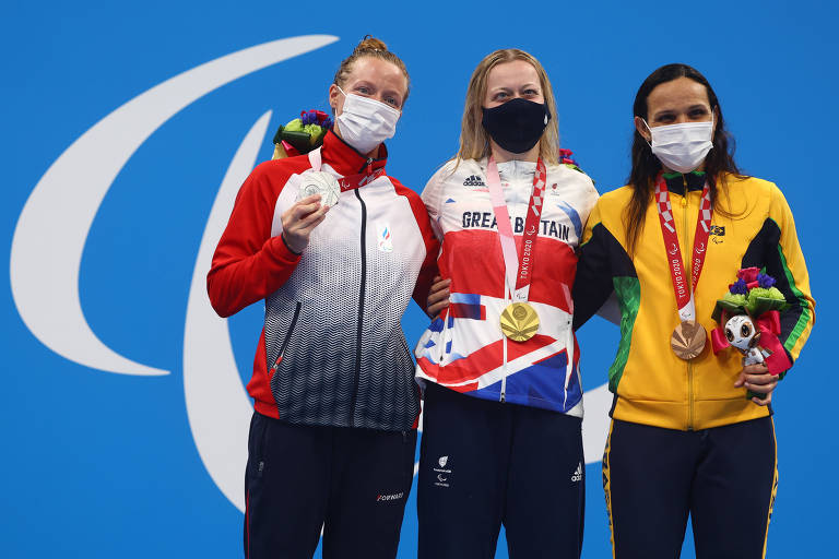 Três atletas abraçadas lateralmente no pódio, todas com medalhas e usando máscaras