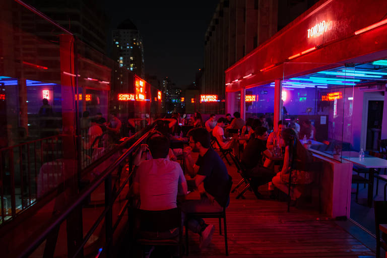 Imagem mostra pessoas sentadas em mesa num bar em São Paulo. O ambiente é iluminado por luzes roxa, vermelha e azul.