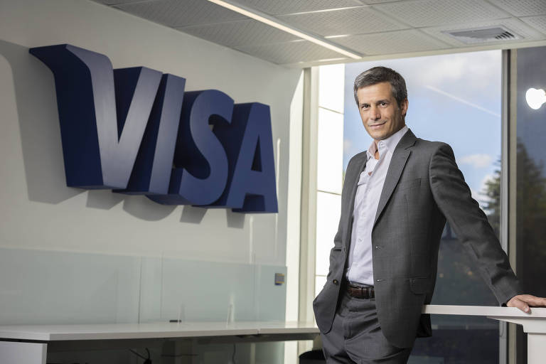 homem branco de cabelo escuro e terno cinza apoiado em mesa em frente ao logo da Visa 