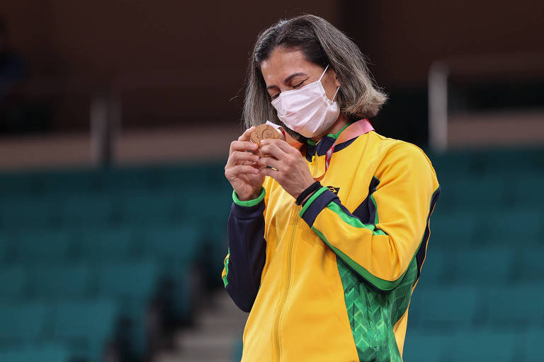 Lúcia Araújo, de agasalho amarelo e máscara, mexe na sua medalha com expressão emocionada