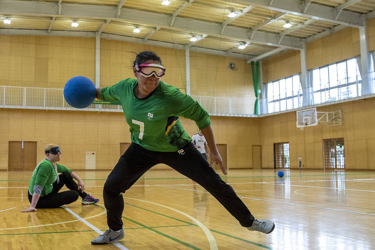 Atleta com camisa verde, calça escura e olhos vendados faz o movimento de lançamento da bola de goalball