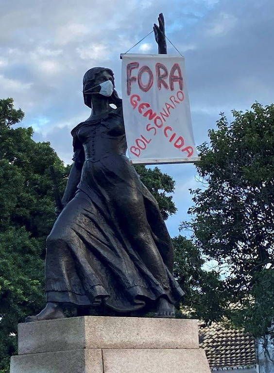 Foto mostra estátua de mulher com placa pendurada em um dos braços; na placa lê-se a frase Fora Bolsonaro genocida