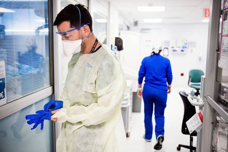 Médico aparece no corredor de um hospital vestindo luvas azuis. Ele usa máscara, jaleco branco e óculos de proteção